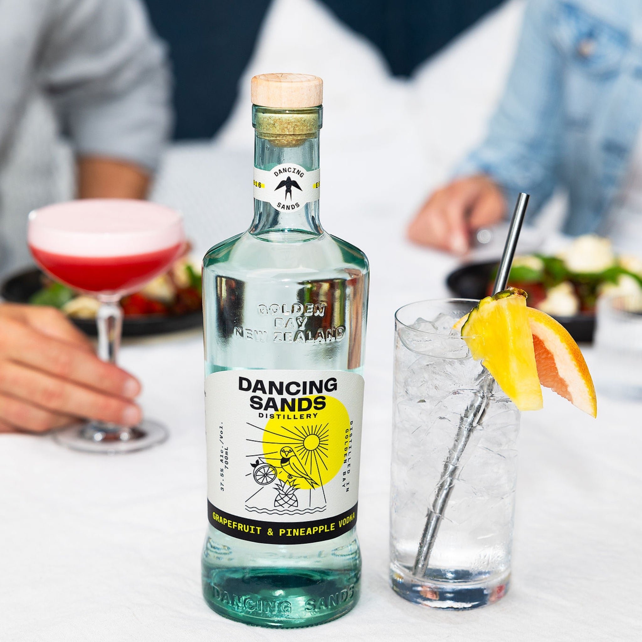 Bottle of Dancing Sands Grapefruit and Pineapple Vodka with cocktails in the background