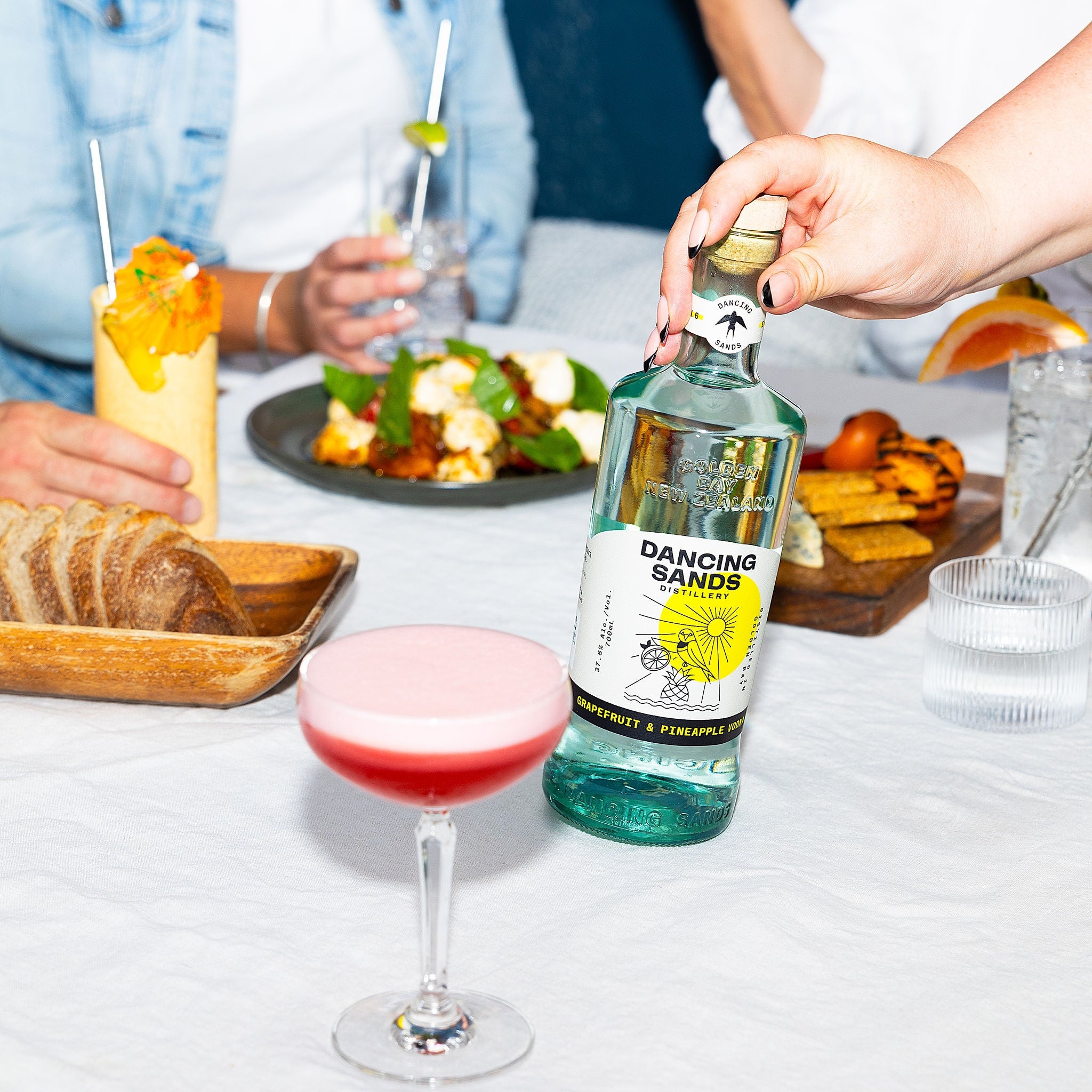 Bottle of Dancing Sands Grapefruit and Pineapple Vodka on a table with cocktails and food