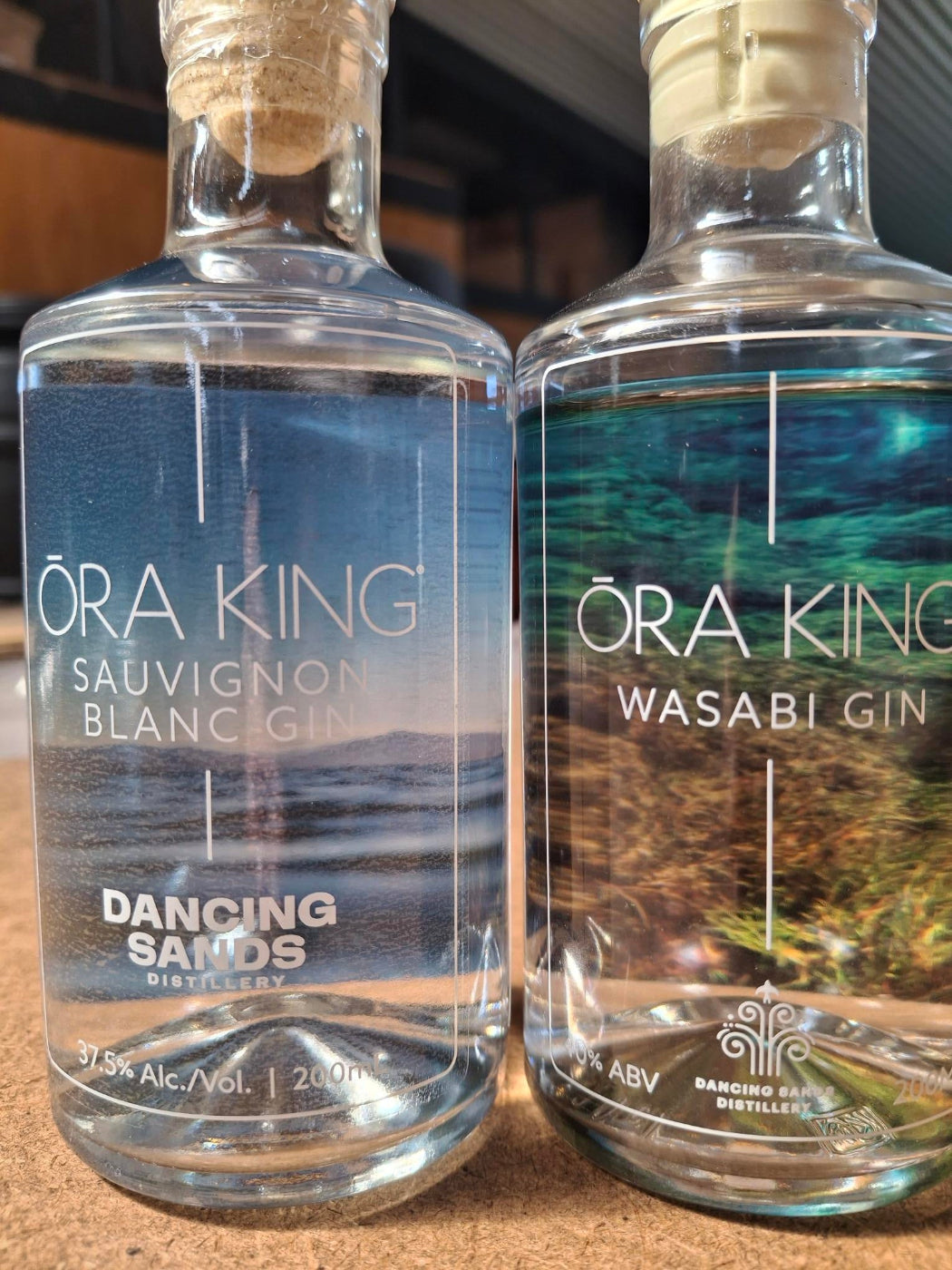 Two bottles of Ora King Gin laid horizontally on a surface, one labeled Sauvignon Blanc Gin and the other Wasabi Gin, with clear glass bottles and cork stoppers visible.