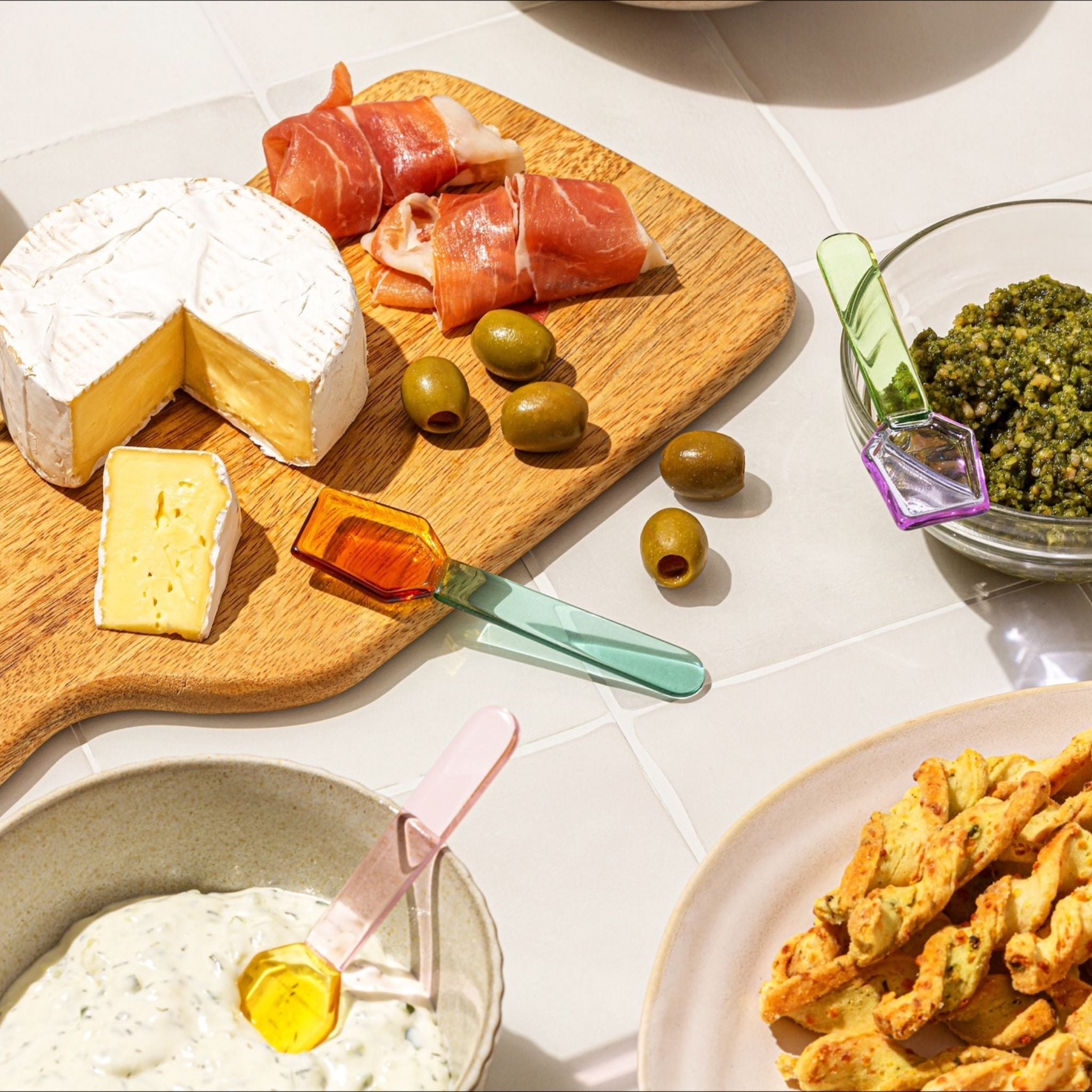 Assorted cheese platter with prosciutto, olives, and a small bowl of green spread on a white surface. Showcasing glass re-useable multicoloured spoons