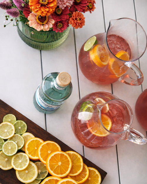 Two pitchers of pink lemonade with oranges and limes on a white wooden table, accompanied by a bottle of Smirnoff Vodka.