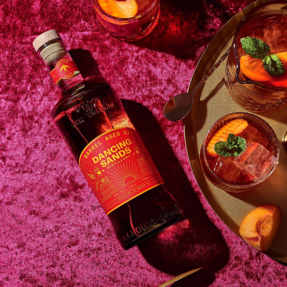 Bottle of Dancing Sands Barrel aged Gin on a pink textured surface with cocktails in the background.