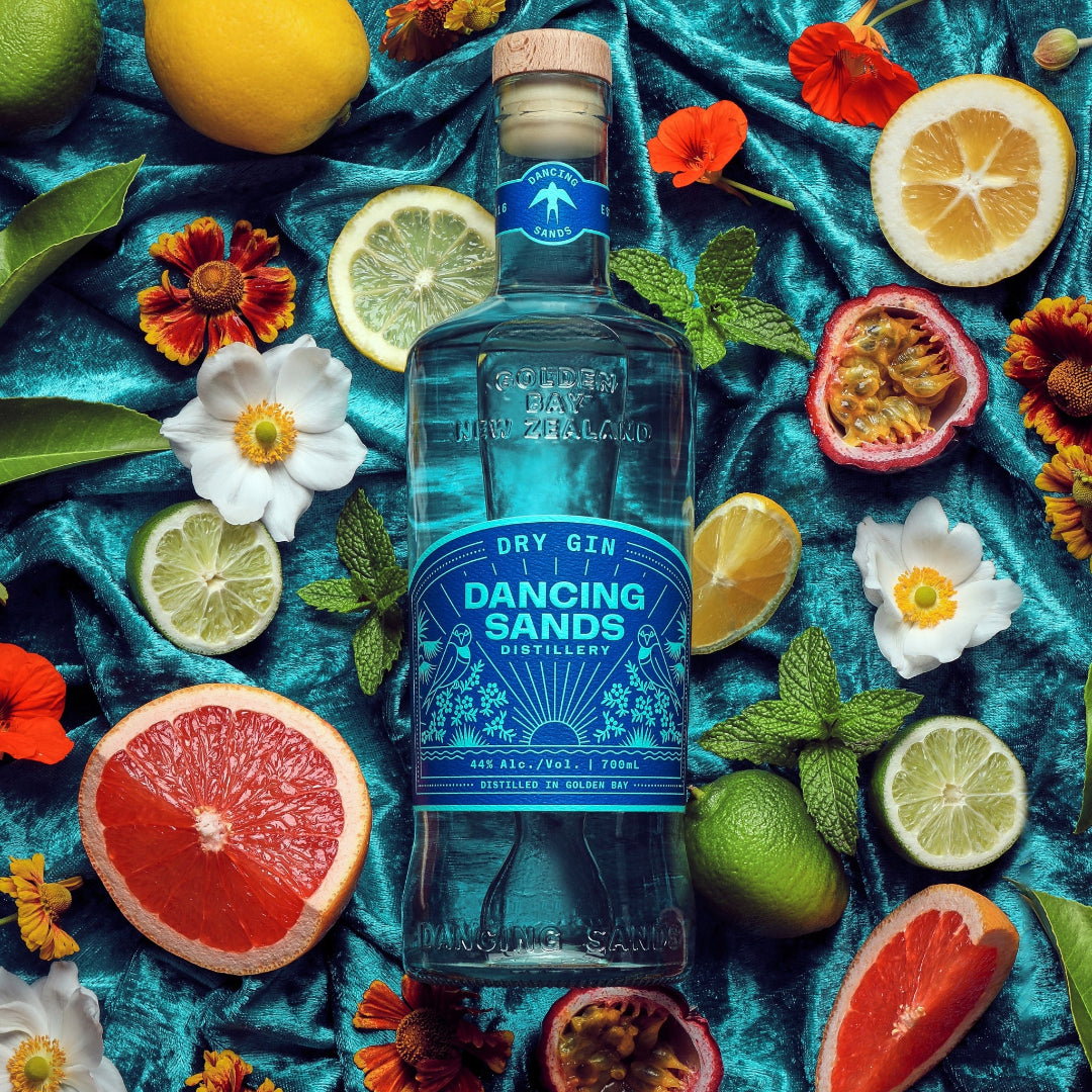 Bottle of Dancing Sands Dry Gin surrounded by citrus fruits and flowers on a blue fabric background