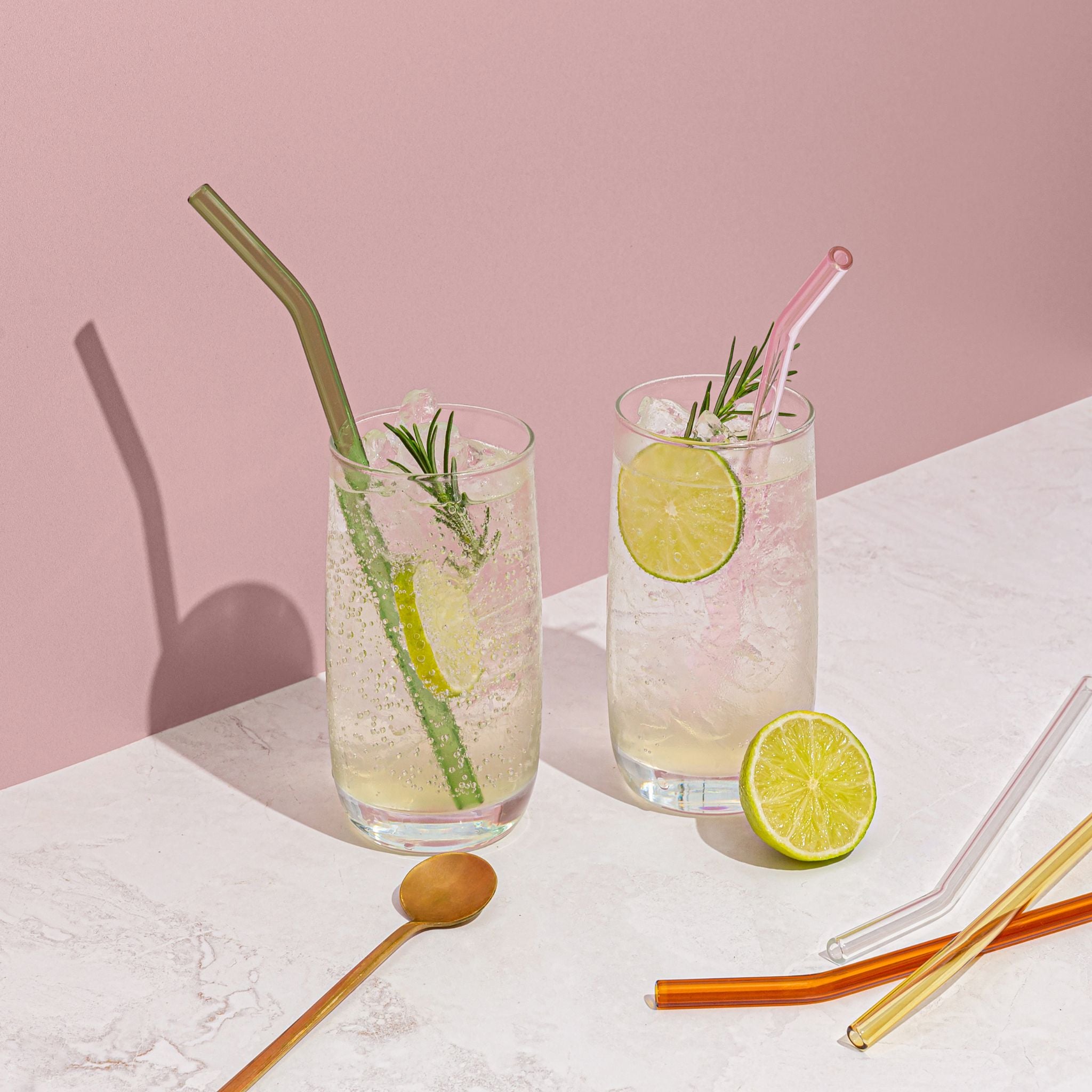 Two glasses of lemonade with ice, lime slices, and rosemary on a pink background.