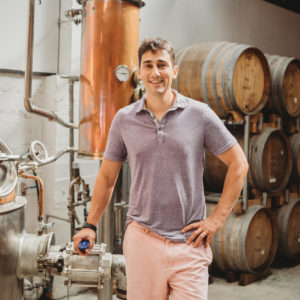 Ben Bonoma Staff Photo In Front Of The Still And Wooden Barrels